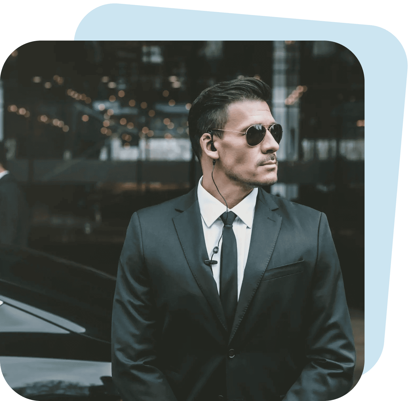 Man in black suit and sunglasses standing confidently outdoors.