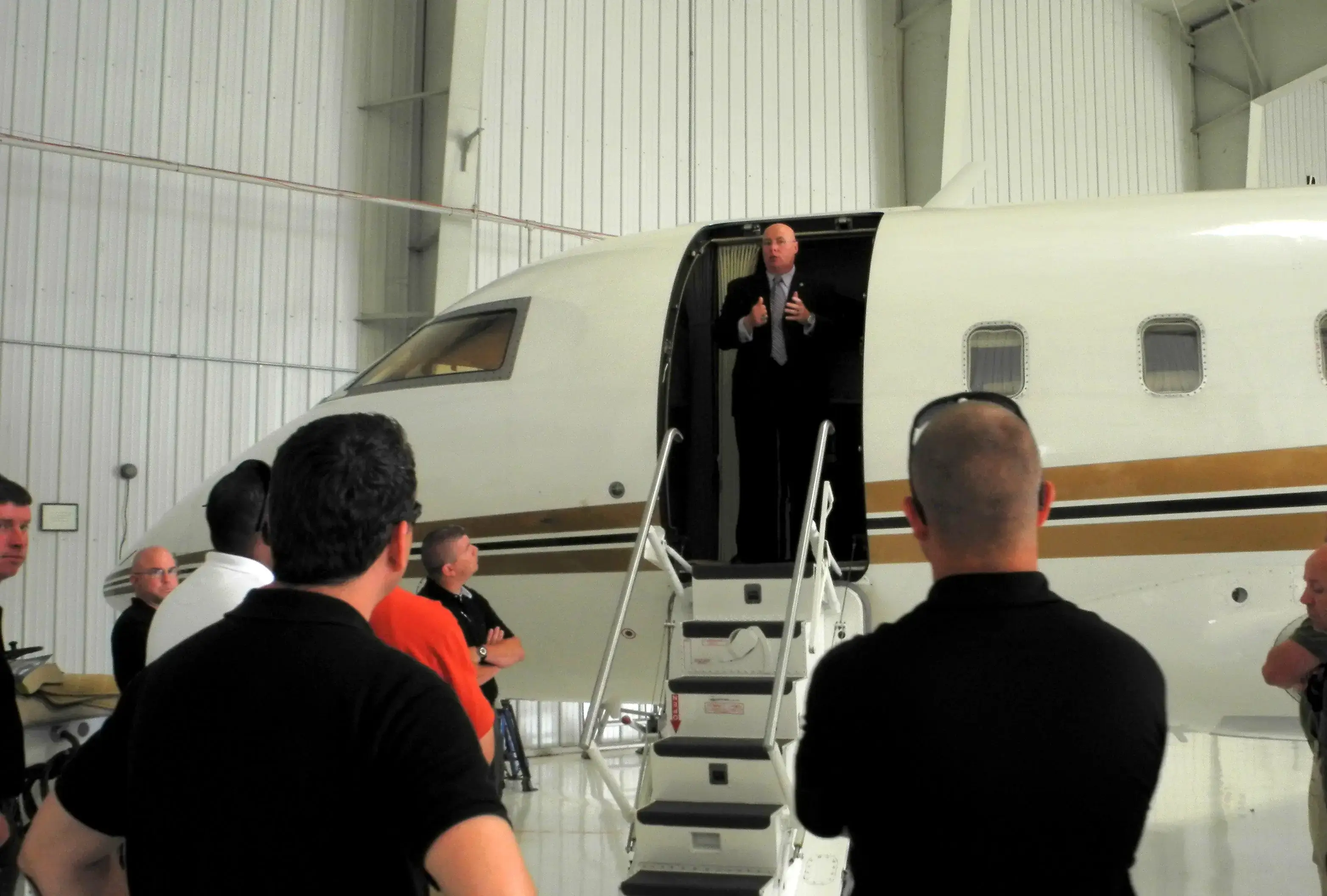 A man in a suit waves from a private jet's doorway to a small crowd.